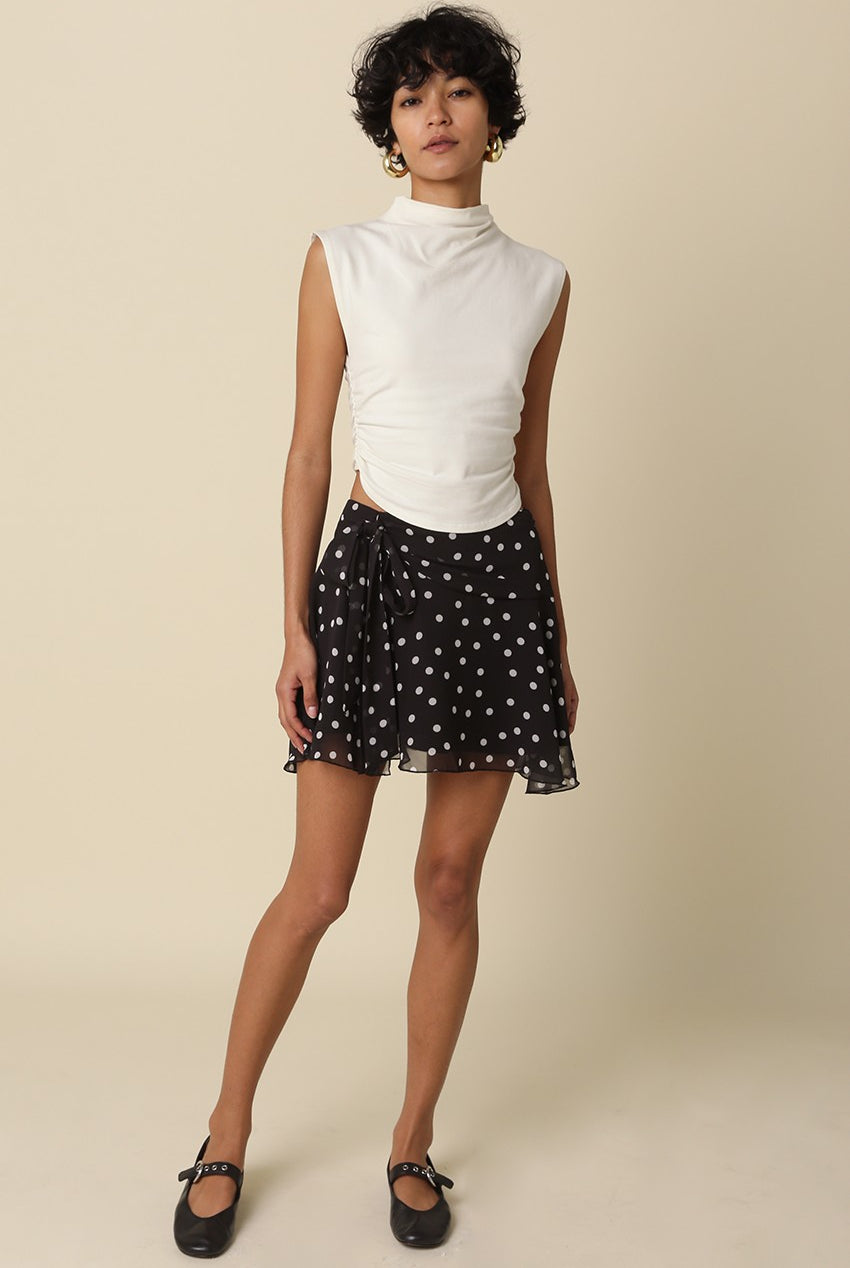 Full length view of a model wearing the Eleni black polka dot ruffle mini skirt with a white mock-neck top and black flats