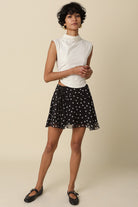 Full body shot of a model styling the Eleni black polka dot ruffle mini skirt with a white sleeveless top