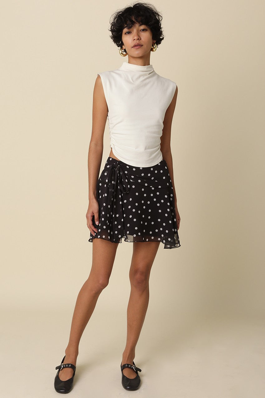 Full length view of a model wearing the Eleni black polka dot ruffle mini skirt with a white mock-neck top and black flats